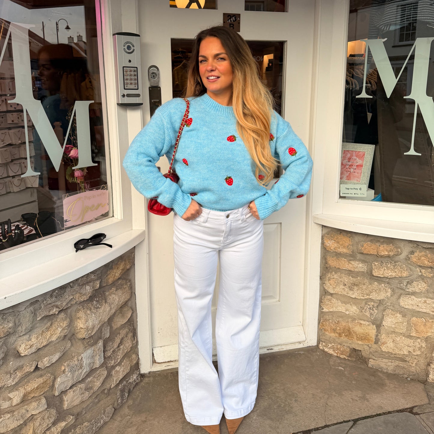 Woman in a blue sweater with red strawberries embroidery and white pants standing in front of a store entrance.