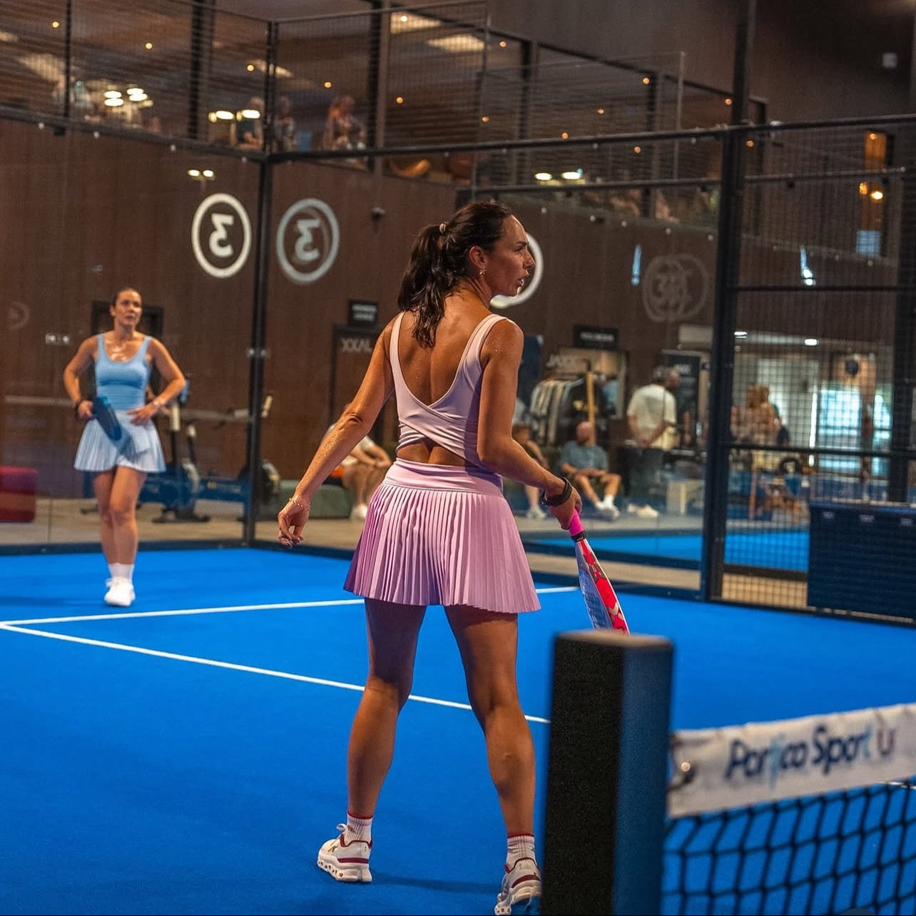 A woman in a pink tennis dress stands on a blue indoor court holding a racket, with another player in a light blue dress in the background. A Periga Sport net is visible in the foreground.