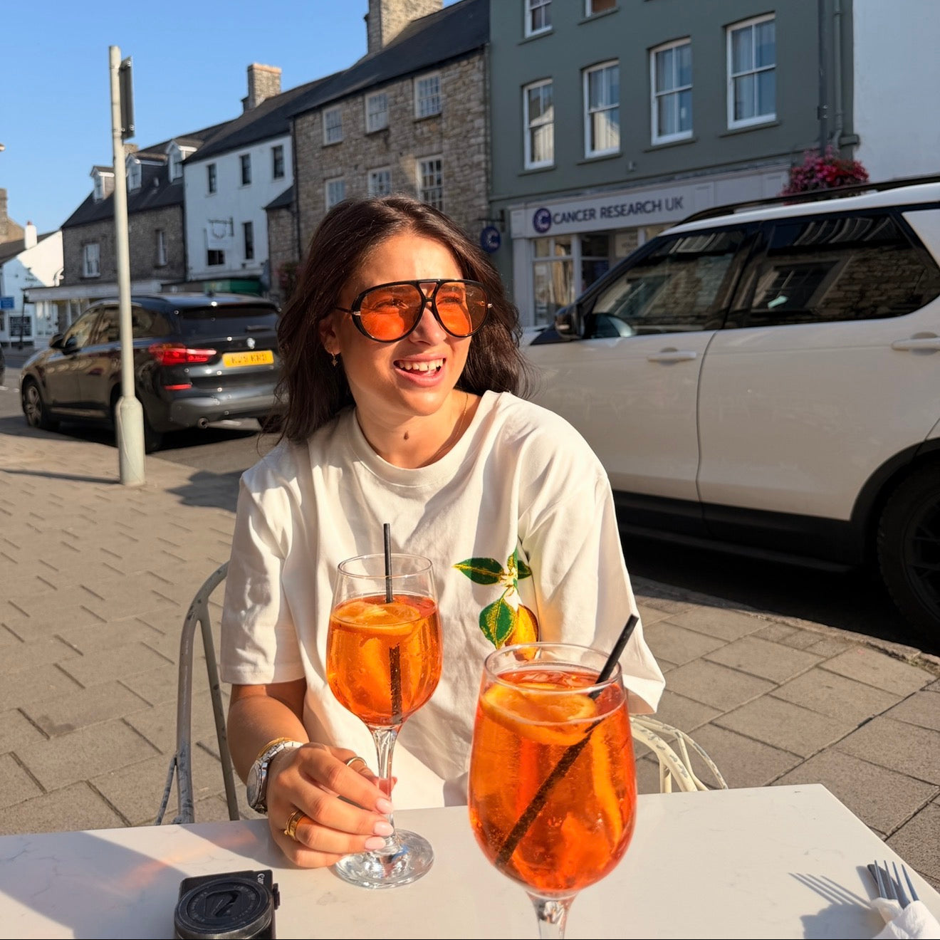 A woman wearing orange-tinted sunglasses and a white graphic t-shirt sits at an outdoor cafe table with two glasses of orange-fruit iced coffee in front of her. The background shows a street scene with parked cars and stone buildings under a clear blue sky.