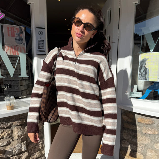 A woman wearing a brown and beige striped half-zip knit sweater and brown leggings stands on a sidewalk in front of a storefront.